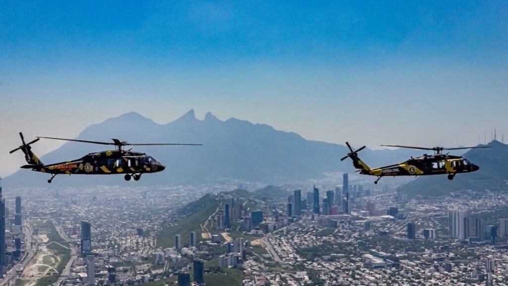 Two Helicopters with Yellow Markings Hover Above a City, Mountains in the Distance Under a Blue Sky - La Razón Nl