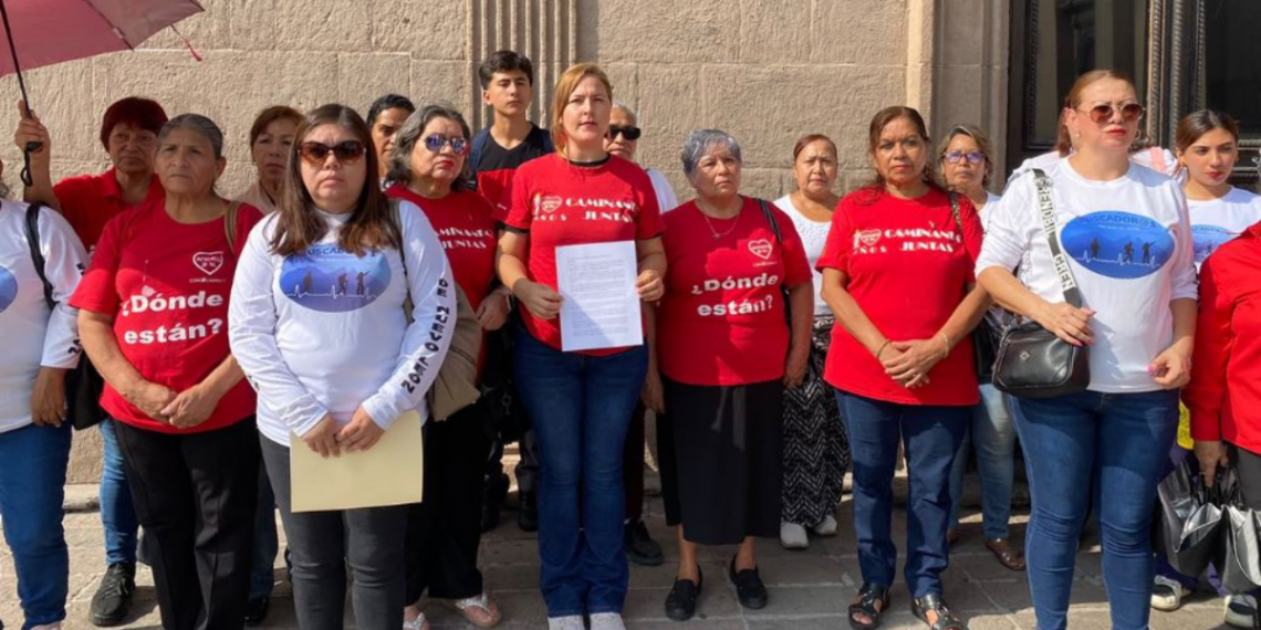 Group of Diverse Adults Standing Together Outside a Stone Building, Many Wearing Red Shirts That Read 'dónde Están?', Some in White Shirts with Blue Graphics, One Person Holding a Sheet of Paper. - La Razón Nl
