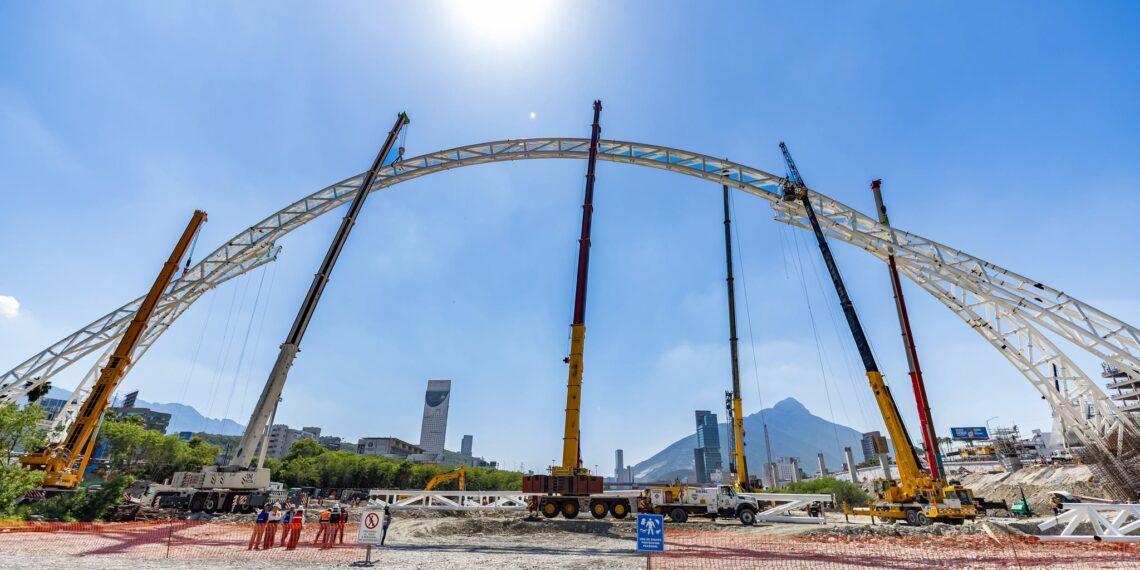 Puente-en-morones-prieto - La Razón Nl Construction Site Showing a Large White Curved Steel Arch Being Lifted by Cranes, with Workers in Orange Vests and Safety Barriers in the Foreground Under a Bright Blue Sky. - La Razón Nl