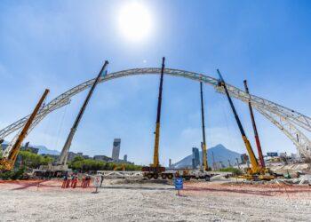 Construction Site Showing a Large White Curved Steel Arch Being Lifted by Cranes, with Workers in Orange Vests and Safety Barriers in the Foreground Under a Bright Blue Sky. - La Razón Nl