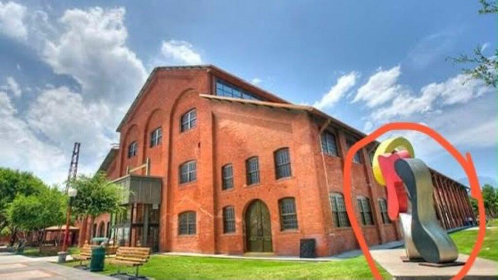 Red Brick Campus Building with a Bright, Abstract Sculpture Featuring Yellow, Red, and Gray Loops in the Foreground on a Sunny Day - La Razón Nl