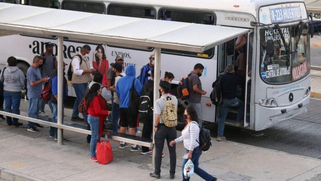 People with Backpacks and Masks Queue at a Bus Stop, Some Boarding a White Passenger Bus. - La Razón Nl