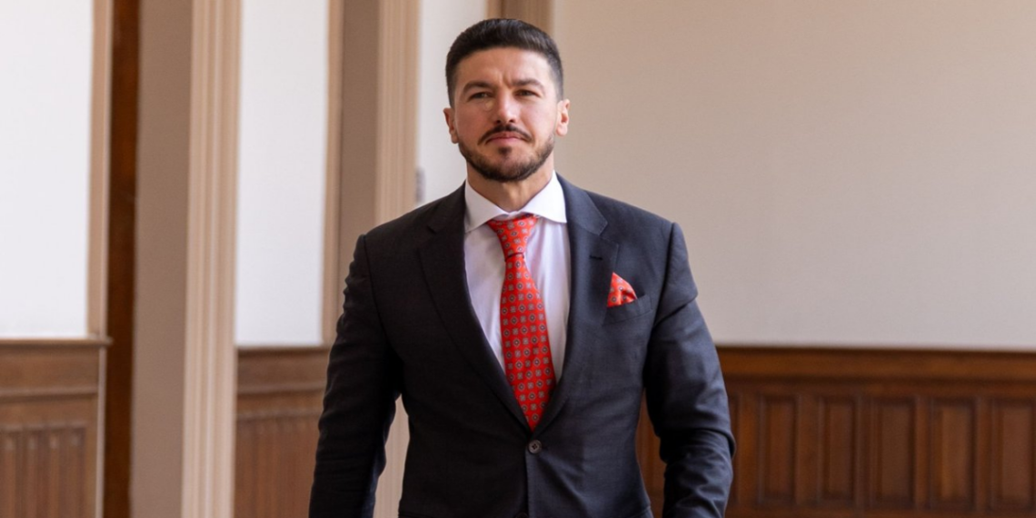 Man in a Dark Suit with a Red Tie and Pocket Square Standing in a Wood-paneled Hallway, Looking at the Camera. - La Razón Nl